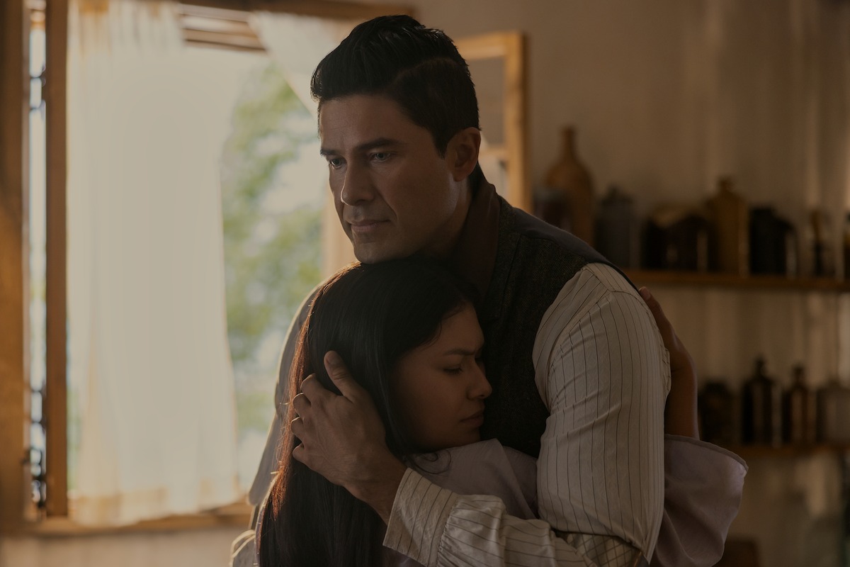 A man and a woman share a comforting embrace inside a warmly lit room with shelves and bottles, sunlight streaming through the window, creating a peaceful, intimate atmosphere.