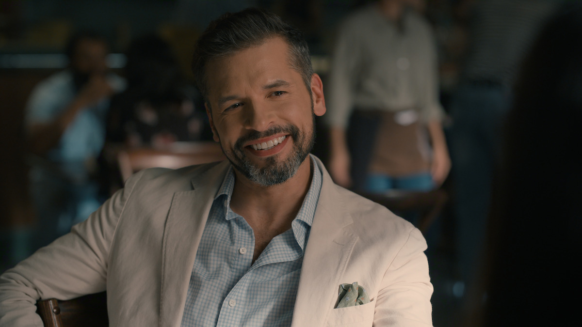 A man with a beard, wearing a light blazer and checked shirt, smiles while sitting at a table in a softly lit café, with blurred people and a casual atmosphere in the background.