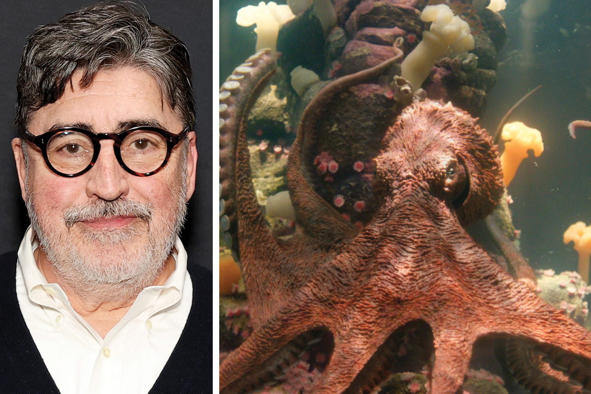 Alfred Molina, with glasses and a beard stands in front of a dark background; next to him, an octopus is underwater among rocks and sea life in an aquarium setting.
