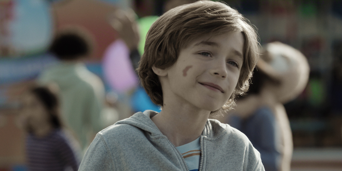 Smiling young boy with a birthmark on his cheek stands outdoors at a fair or carnival, wearing a gray hoodie, with blurred people and colorful decorations in the background.