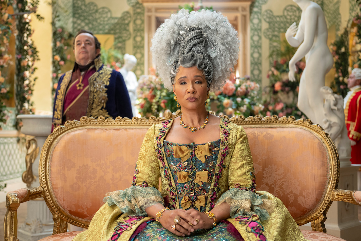 Woman in elaborate 18th-century dress and white wig sits on ornate sofa in a lushly decorated palace room, with a man in period costume standing behind her and floral arrangements in the background.