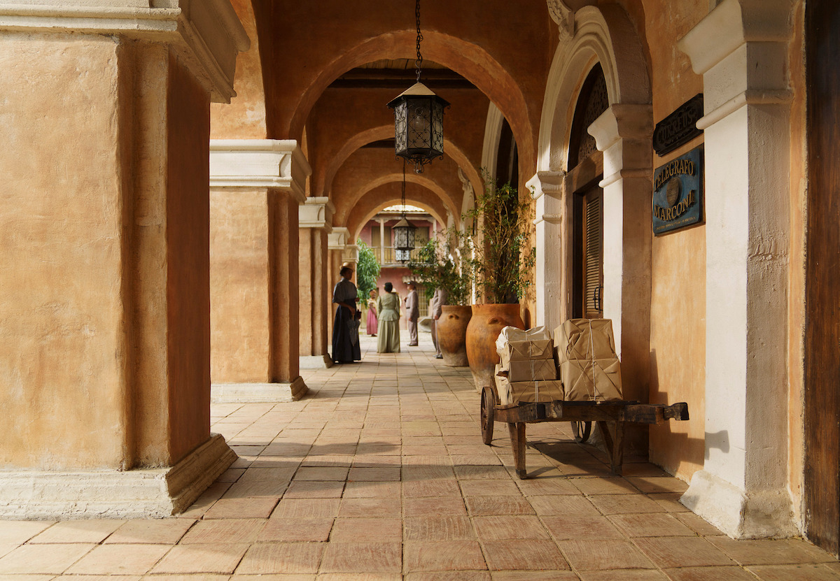 Old stone hallway with arches, a lantern, plants, large clay pots, a wooden cart stacked with boxes in the foreground, and people in historical clothing standing in the background. Warm, sunlit setting.