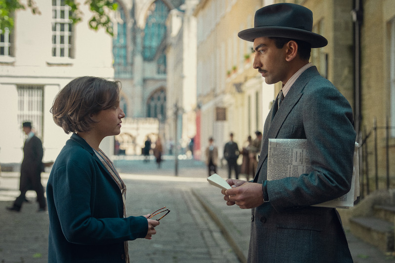 Two people in period clothing stand on a cobblestone street in a historic city, engaged in a serious conversation; the atmosphere is tense and thoughtful, with soft daylight and blurred figures in the background.
