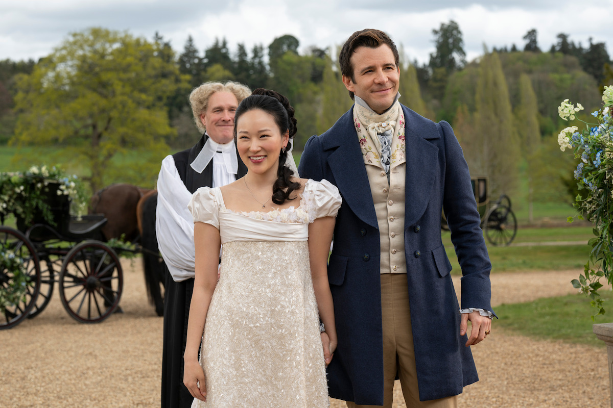 A smiling couple in elegant Regency-era attire stands outdoors on a gravel path, holding hands, with a carriage, greenery, and a formally dressed man in the background, evoking a historical or period drama setting.