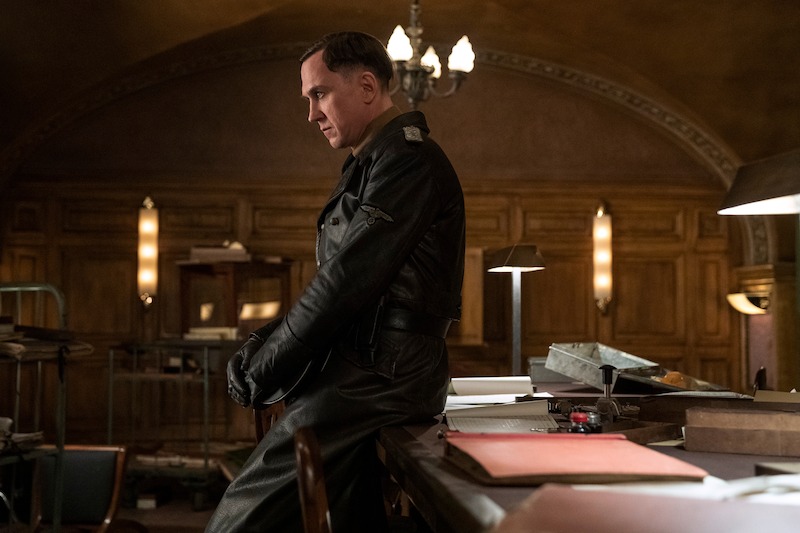 Lars Eidinger as Sergeant Major Reinhold von Rumpel sitting on the edge of a desk, looking down. 