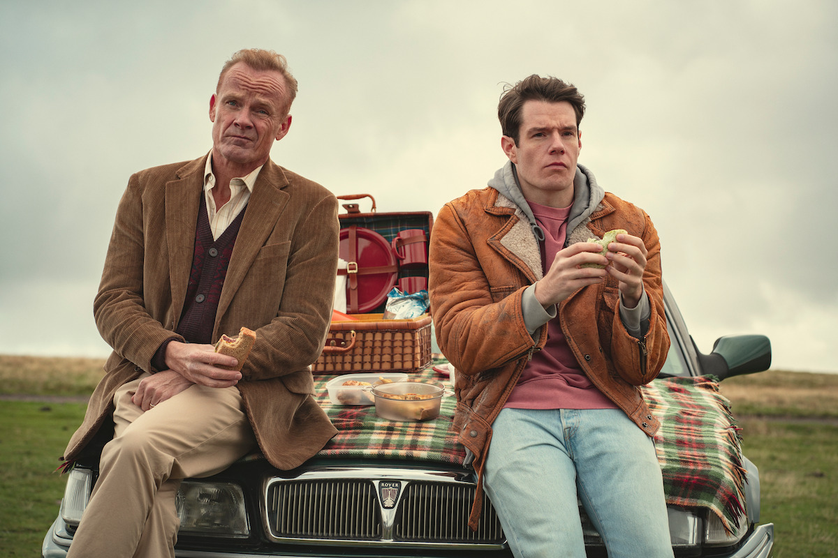Alistair Petrie as Michael and Connor Swindells as Adam sit on the hood of a car together in Season 4 of ‘Sex Education.’