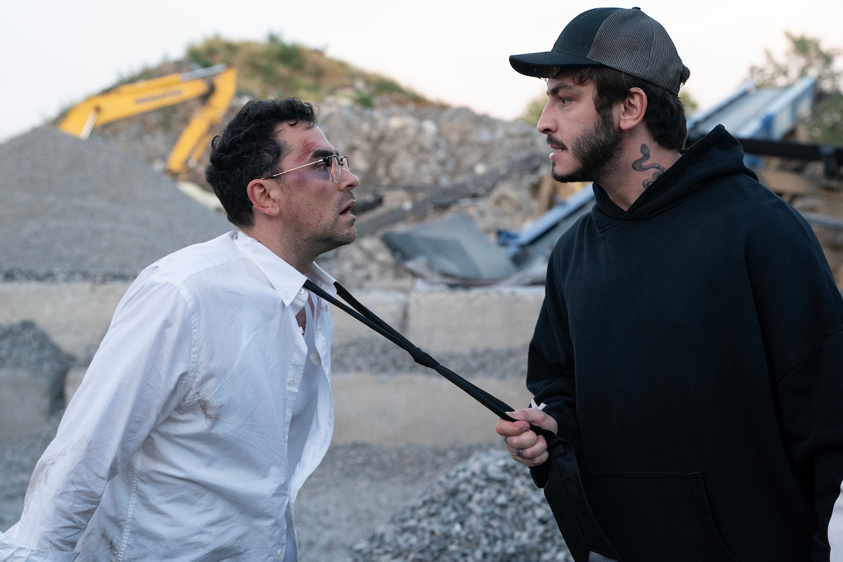 Two men confront each other at a construction site with gravel piles and machinery; one man in a white shirt appears injured and is being grabbed by the collar by the other man wearing a black hoodie and cap.