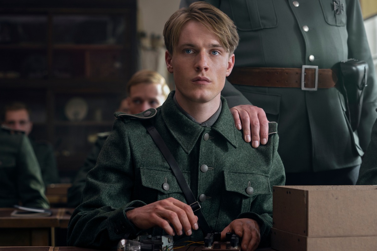 Werner Pfennig (Louis Hofmann), a teenage boy with light features, dirty-blonde hair and blue eyes sits at a desk in a military uniform. A military officer has his hand on Werner’s shoulder and there are other young military recruits in the background. On the desk in front of Werner sits an assembled radio and cardboard box.