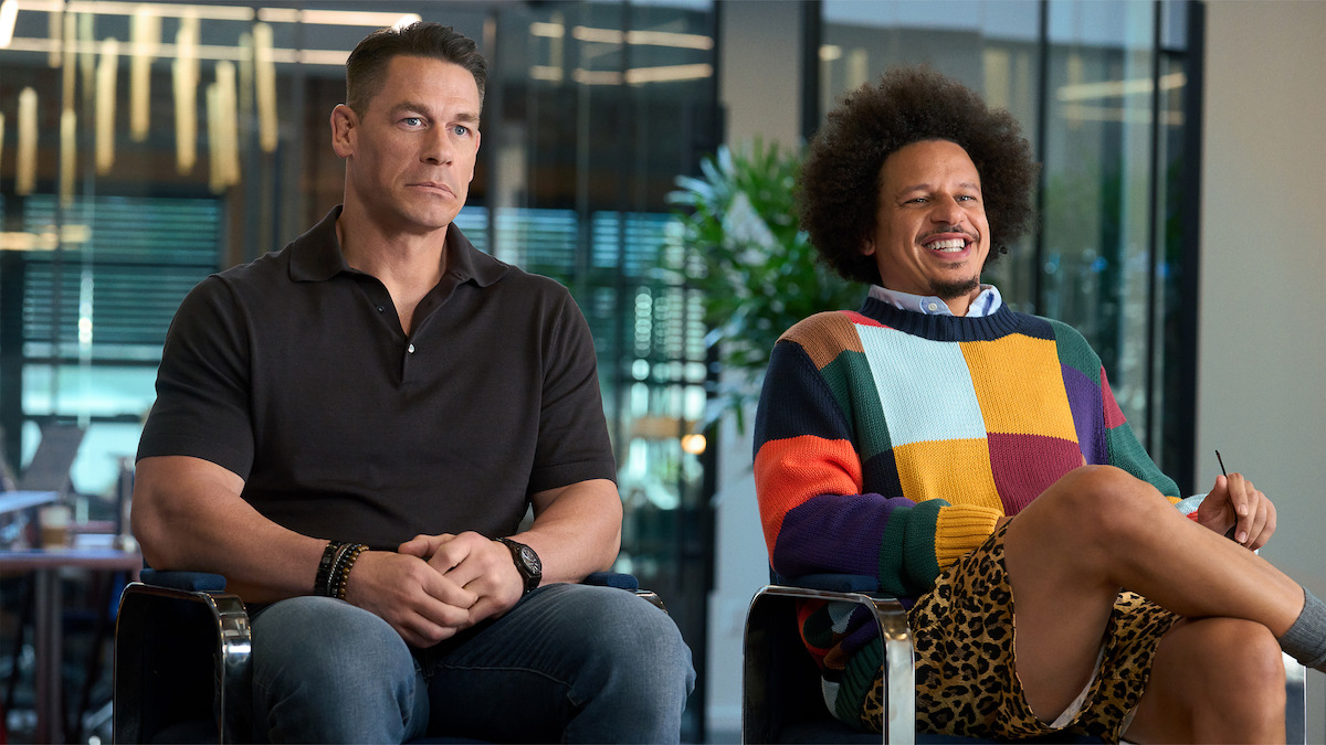 Two men sitting indoors on office chairs; one wears a black polo and jeans looking serious, the other wears a colorful sweater and shorts, smiling, with greenery and modern office decor in the background.