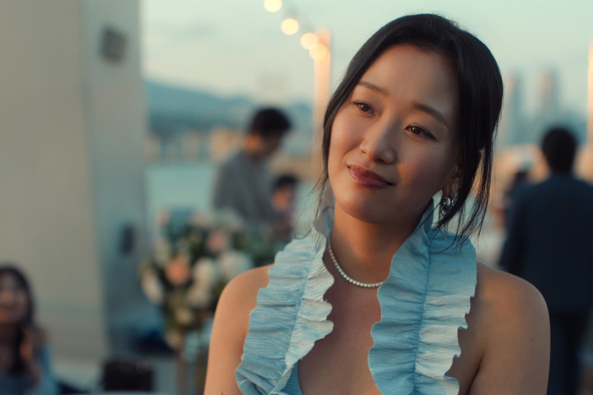 Woman in a light blue ruffled dress and pearl necklace at an elegant outdoor event, with blurred people, floral arrangements, and cityscape in the background during dusk.