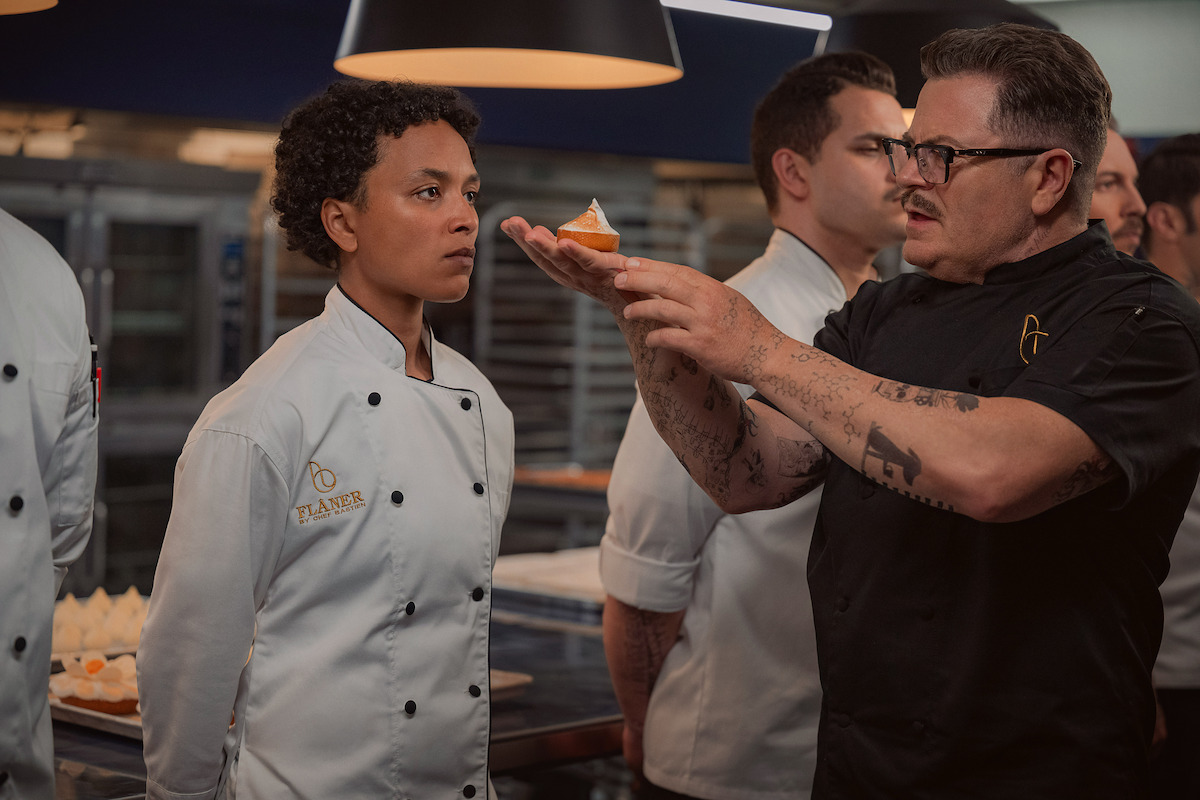 Chef in a black coat explaining a plated dessert to a focused chef in a white coat in a professional kitchen, with other chefs and kitchen equipment visible in the background.