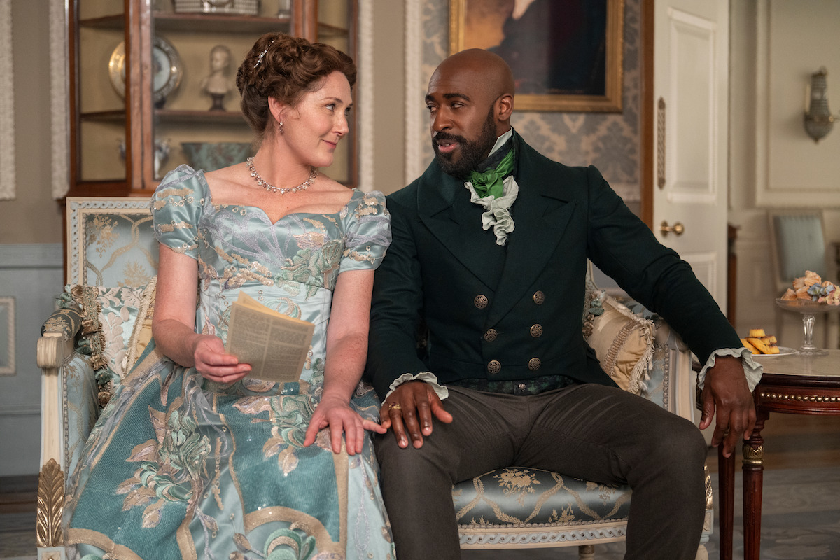 A woman in a floral Regency dress holding a book sits on a sofa beside a man in period attire in an elegant, classic room with ornate decor and antique furnishings, both smiling and engaged in conversation.