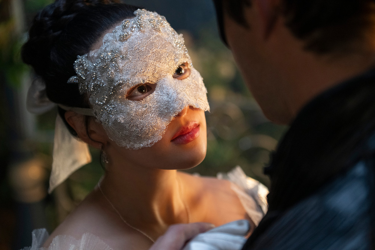 A woman in an ornate lace mask gazes intently at a man in a dim, romantic setting, suggesting a mysterious and intimate moment at a masquerade or costume event.