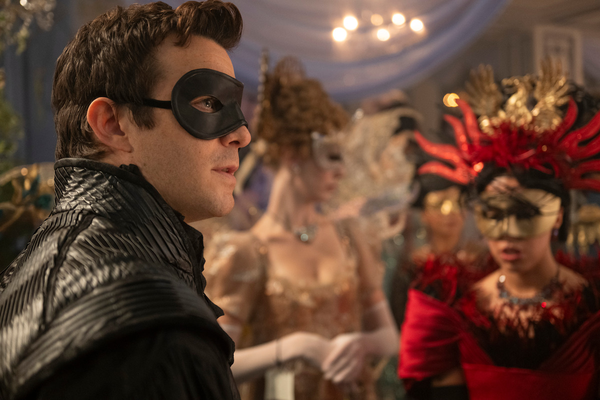 A man in a black mask and costume at a lavish masquerade ball, surrounded by other elegantly dressed guests wearing ornate masks and formal attire in a decorated, chandelier-lit room.