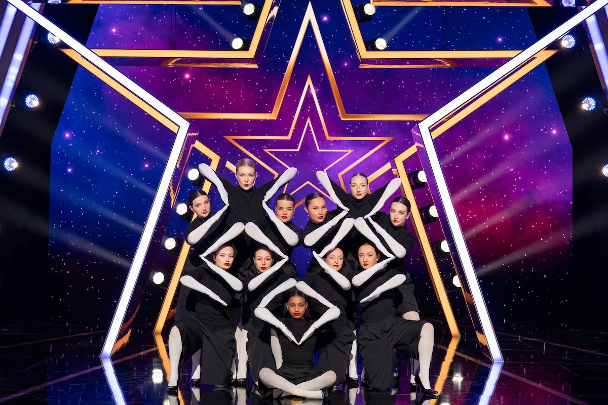 Ladymetry in black and white costumes performs a synchronized formation on a stage with a large illuminated star backdrop and vibrant purple and blue lighting effects.