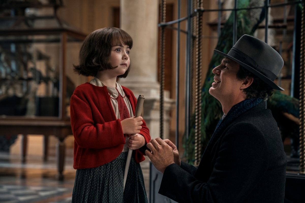 Young Marie-Laure (played by Nell Sutton) stands in the aviary of a museum with Daniel LeBlanc (played by Mark Ruffalo) who is crouched down in front of her and smiling. Marie-Laure is holding a white cane and wearing a red cardigan and blue skirt. Daniel is wearing a dark coat and hat.