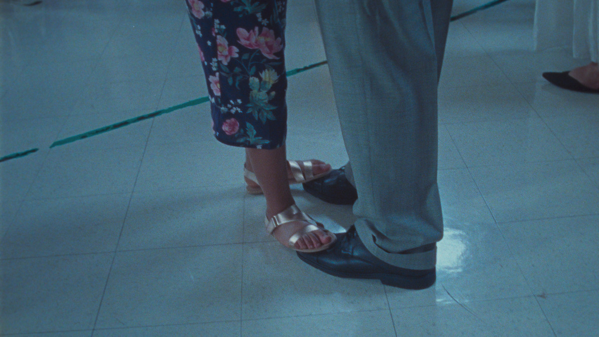 Gold-sandaled feet stand atop black dress shoes.