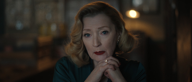 A woman with styled blonde hair and red lipstick sits with her hands clasped under her chin, looking thoughtfully ahead in a softly lit room.