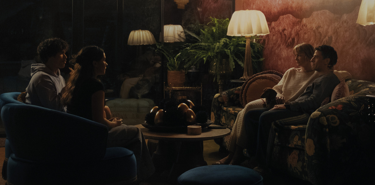 Four people sit in pairs on sofas in a dimly lit, cozy living room with lamps, houseplants, and a central table, creating a warm, intimate atmosphere.