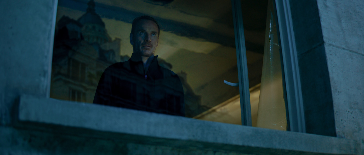 Michael Fassbender as The Killer wears all black and looks through a window in ‘The Killer’