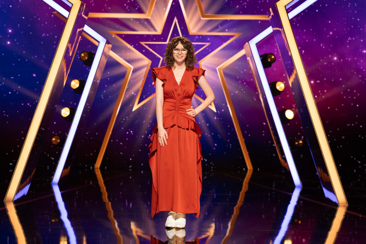 Sophie Saylor stands confidently on a glossy stage with bright lights and a large illuminated star in the background, wearing a sheer black and gold dress for a talent or competition show.