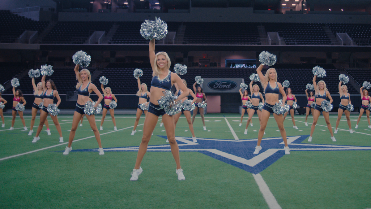 A group of cheerleaders in blue uniforms performing a synchronized routine with pom-poms on a football field inside a large stadium, with audience seating visible in the background.
