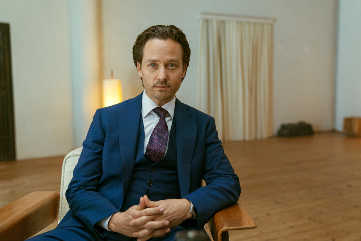 Tom Schilling as Björn in ‘Murder Mindfully’ wears a navy three-piece suit and smugly looks into the camera while he sits on a chair with his hands interlocked.