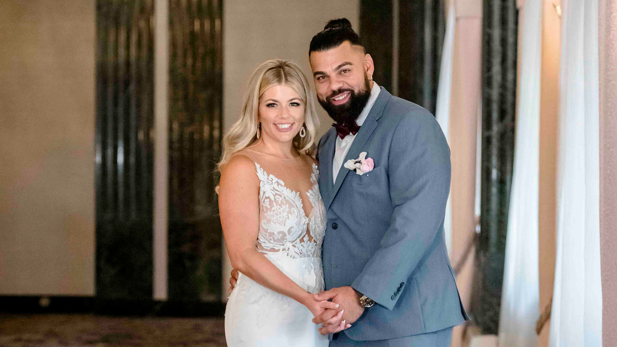 Bride in white dress and groom in blue suit posing together indoors, holding hands and smiling, elegant setting with soft lighting and decorative background elements.