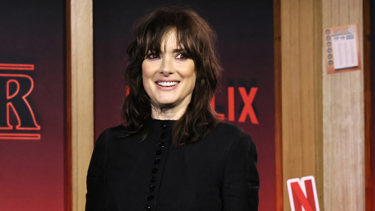 Winona Ryder, dressed in a black outfit, smiles at an indoor Netflix event, posed against a backdrop featuring red lighting and Netflix branding that creates an energetic, celebratory atmosphere.