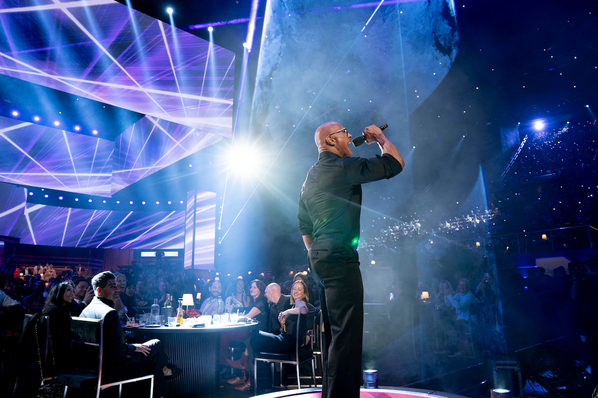 A singer performs on stage under bright lights and fog effects at a crowded indoor concert or awards show, with an audience seated at tables and colorful laser lighting in the background.