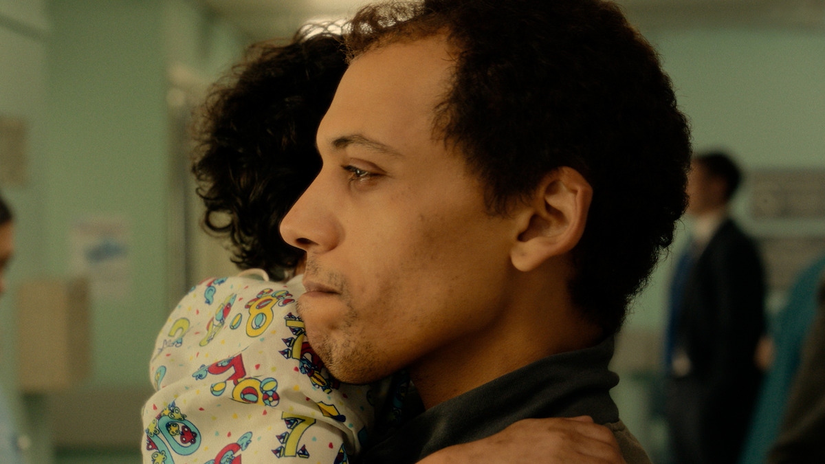 A man in a hospital gently holds a child in colorful pajamas, looking concerned. The background shows a medical setting with people and clinical lighting, suggesting an emotional moment in a healthcare environment.