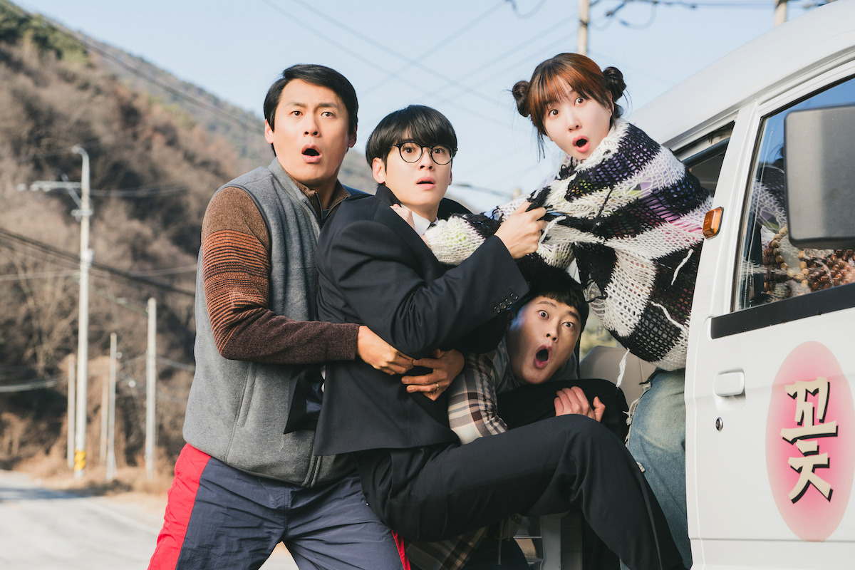 Four surprised young adults stand outdoors by a white van on a rural road, mountains and utility poles in the background, all displaying expressions of shock or alarm, suggesting an unexpected or dramatic event.