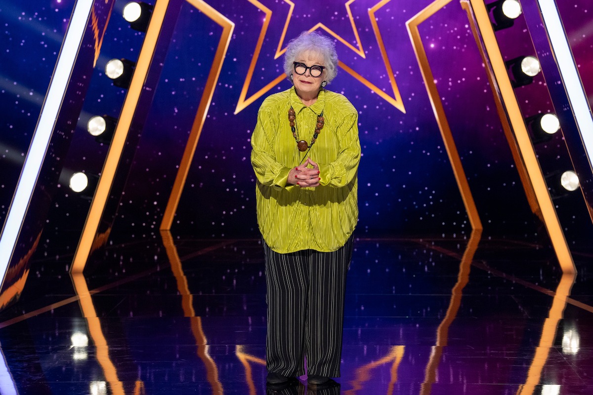 Susan Rice with white hair and glasses stands on a shiny stage with bright lights, wearing a yellow blouse, striped pants, and a necklace. Background features a large glowing star and purple starry pattern.