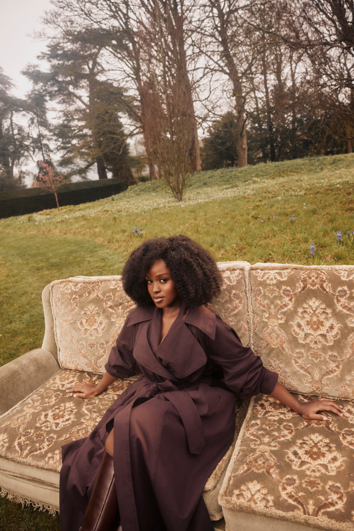 Woman in a brown coat sitting on an ornate vintage couch outdoors in a grassy garden with trees and overcast sky in the background.