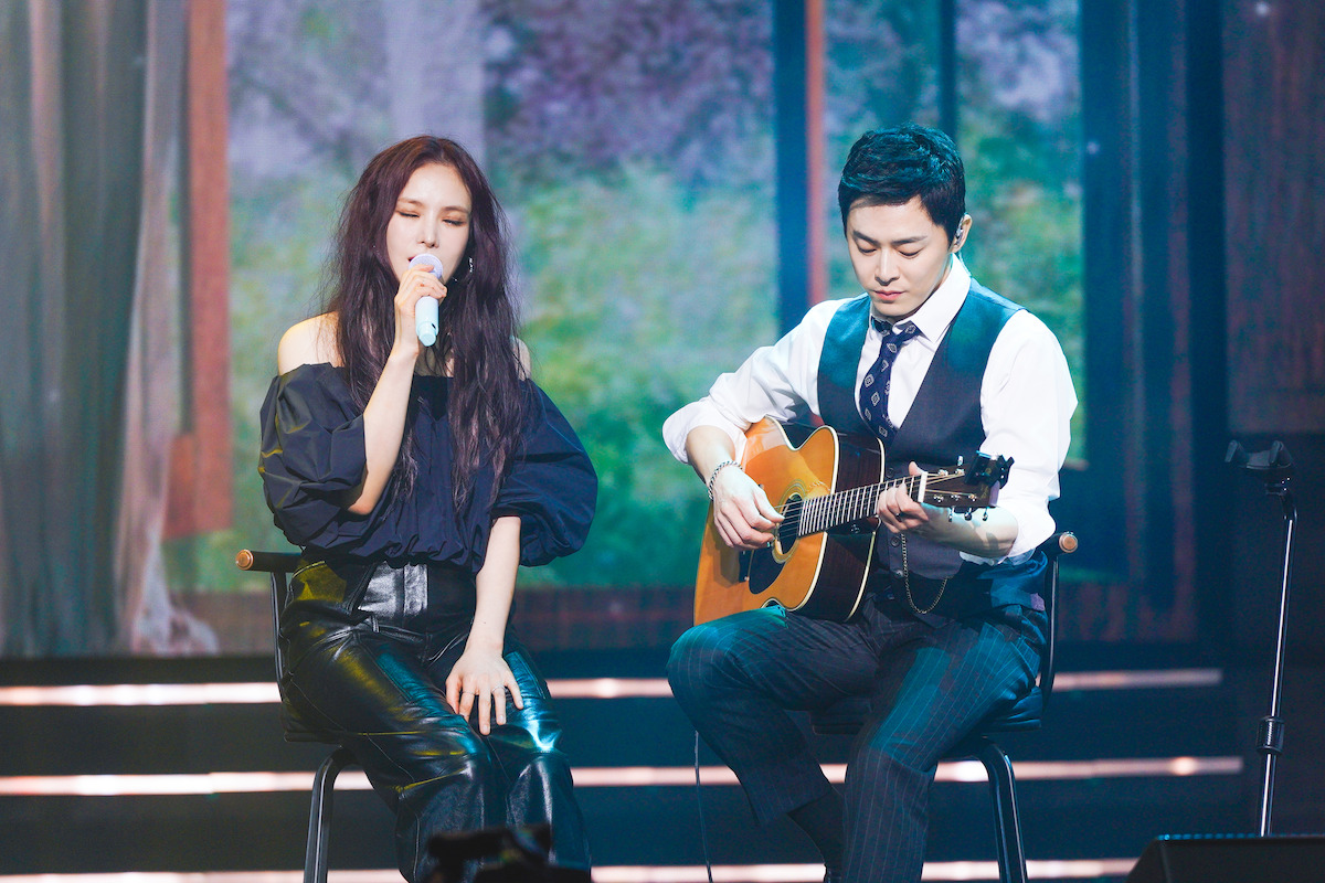 A woman sings while a man plays a guitar on stage in 'A-List to Playlist'.