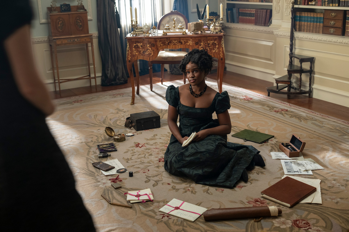 A woman in a dark vintage dress sits on a rug surrounded by letters, books, and papers in an elegant, historical study with ornate furniture and antique decor.