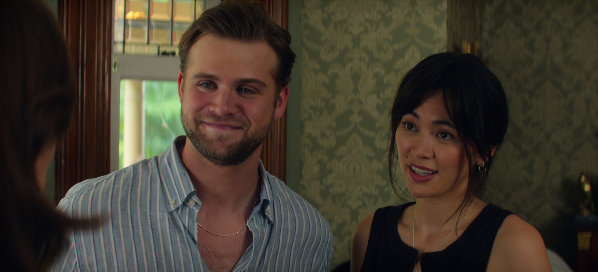 Leo Woodall as Vladimir and Jessica Henwick as Cynthia standing indoors, smiling and talking to another person, with patterned wallpaper and a window in the background, casual atmosphere.