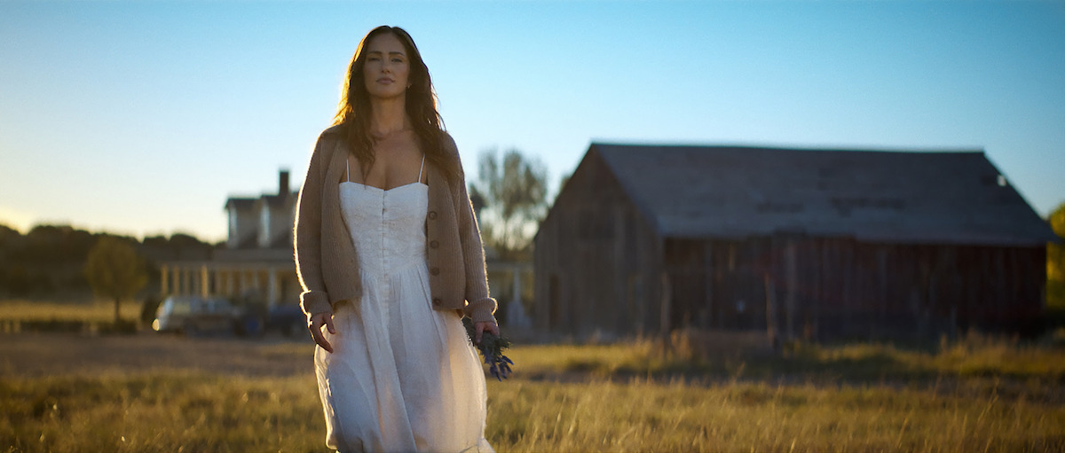 Woman in a white dress and cardigan walks through a grassy field at sunset, holding flowers, with an old barn and farmhouse in the background under a clear blue sky.