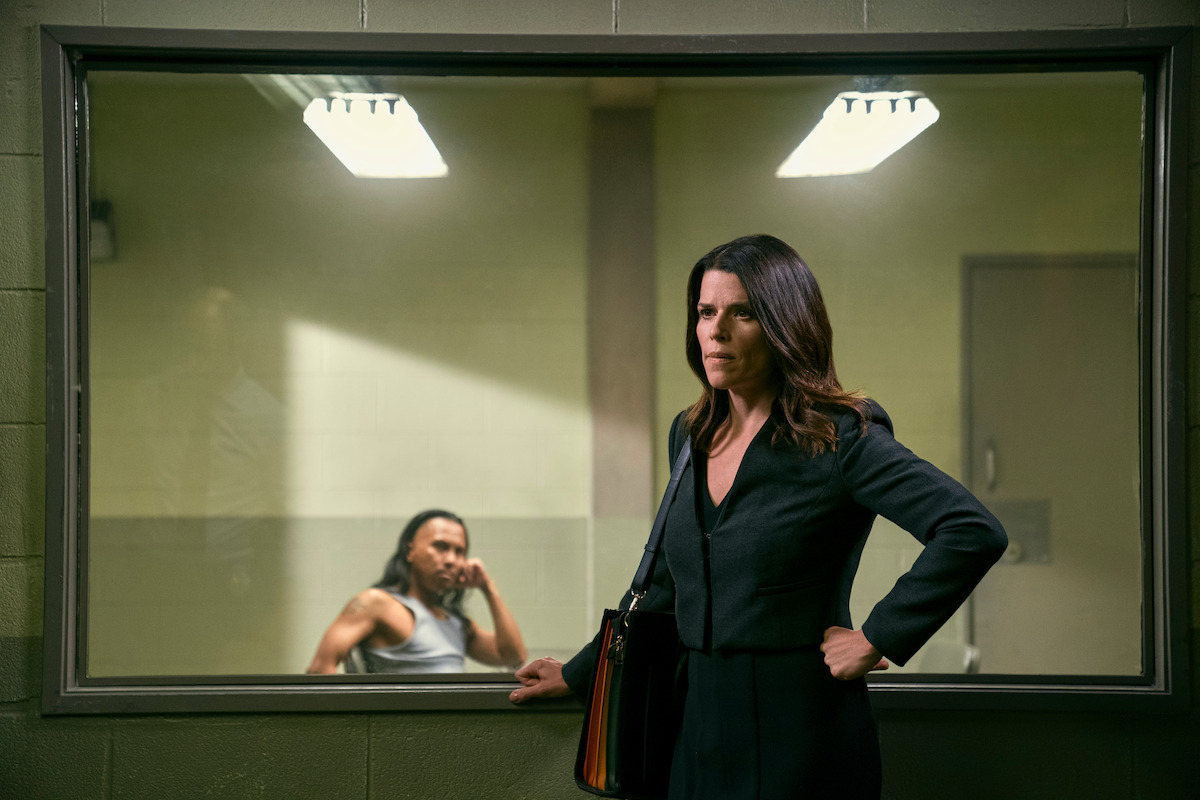 A woman stands in an interrogation room as someone out of focus behind her looks on.