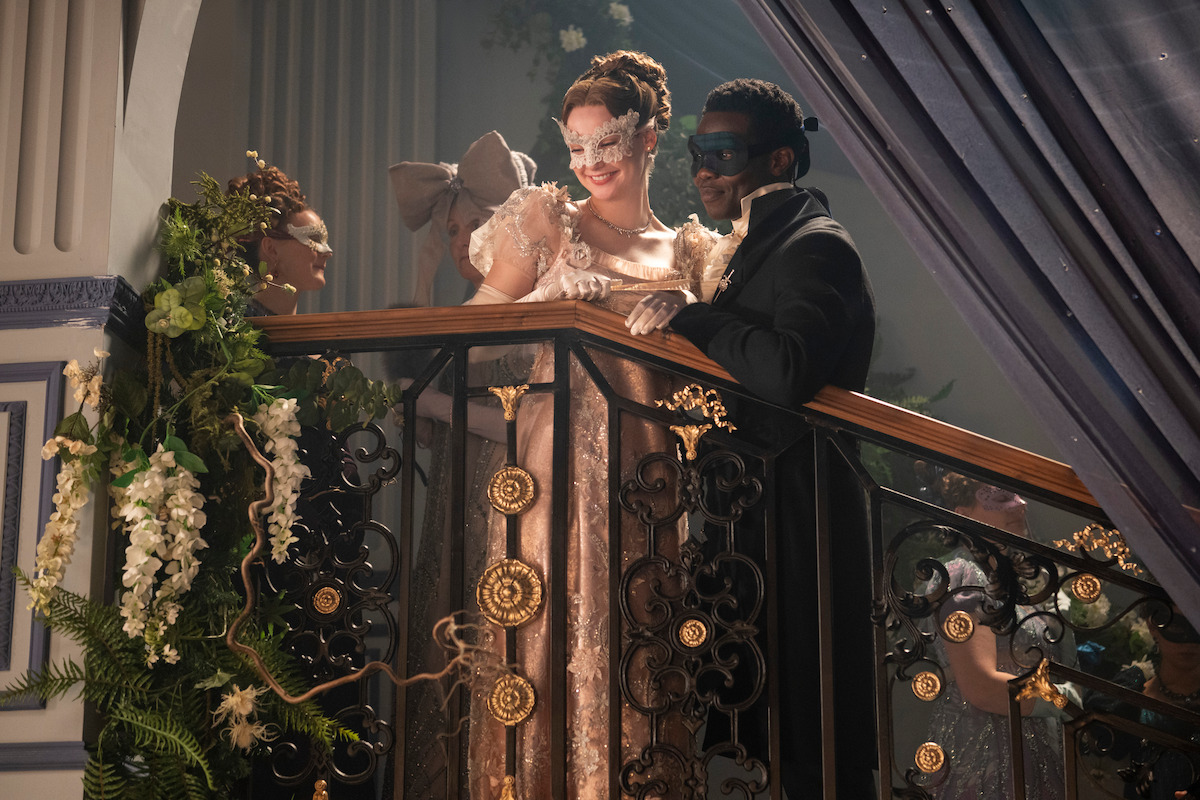 Three people in elegant masquerade attire stand on an ornate balcony decorated with flowers at a lavish indoor event, smiling and conversing under soft lighting.