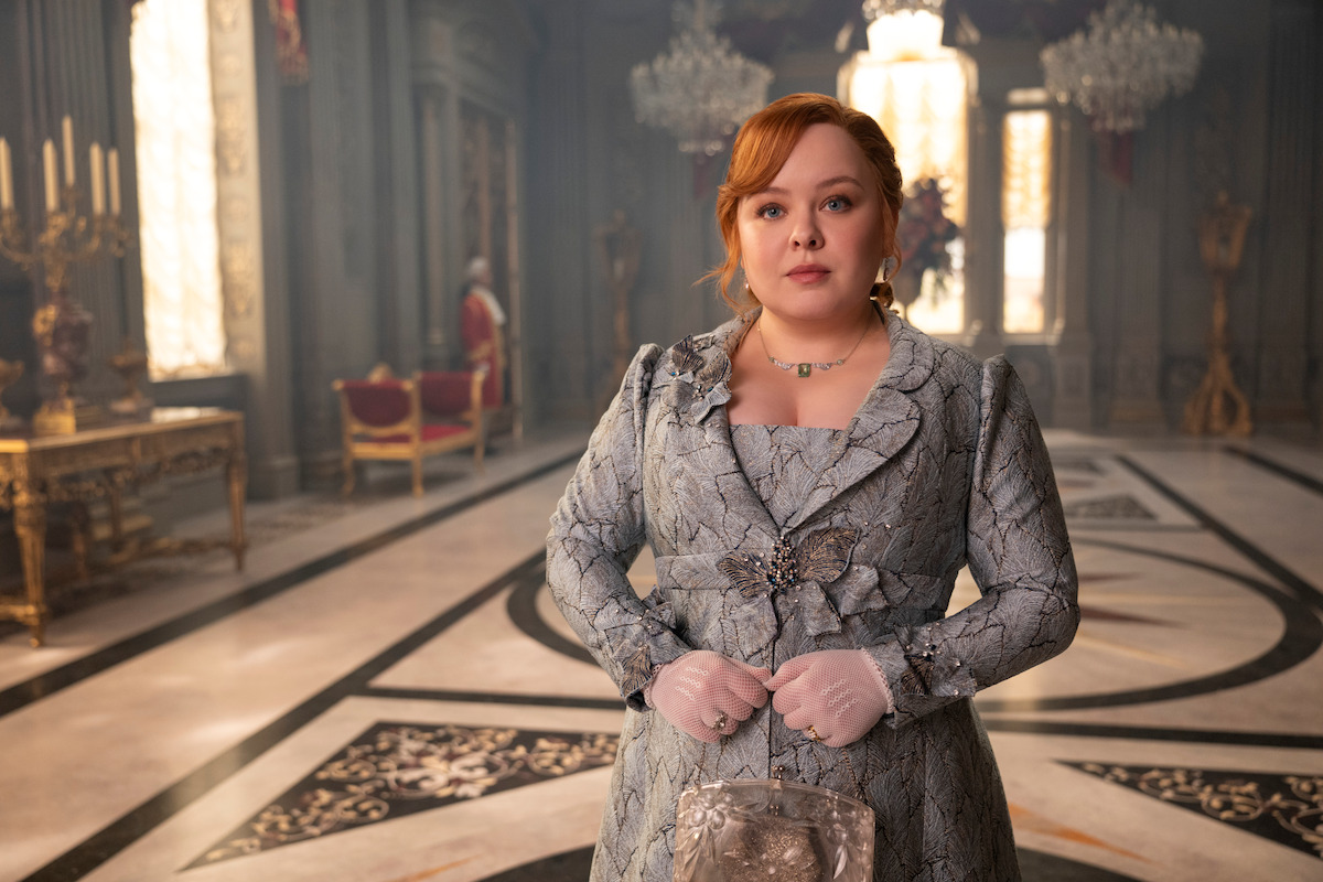 Woman in ornate period costume stands in an elegant palace room with chandeliers, marble floors, and luxurious decor. She looks forward calmly, hands clasped with gloves, and intricate patterns are visible on her dress.
