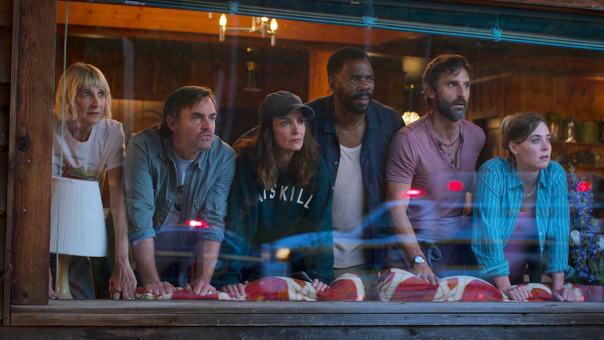 Six people lean on a windowsill inside a rustic wooden building, looking outside with serious, focused expressions. The setting has warm lighting and cozy decor, with reflections of lights and cars visible on the glass.