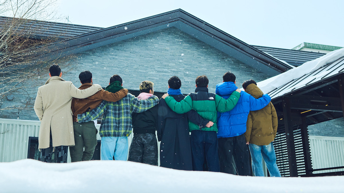 A group of eight people stand arm-in-arm, facing a snow-covered house, dressed warmly in winter clothing. The environment is wintry and serene, conveying friendship and togetherness in a peaceful outdoor setting.