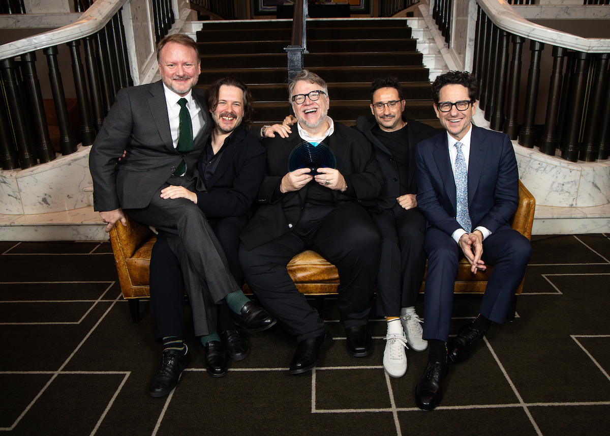 Five men in suits sitting on a gold sofa in front of marble stairs, posing together and smiling in a formal indoor setting. One man in the center holds a round glass award.