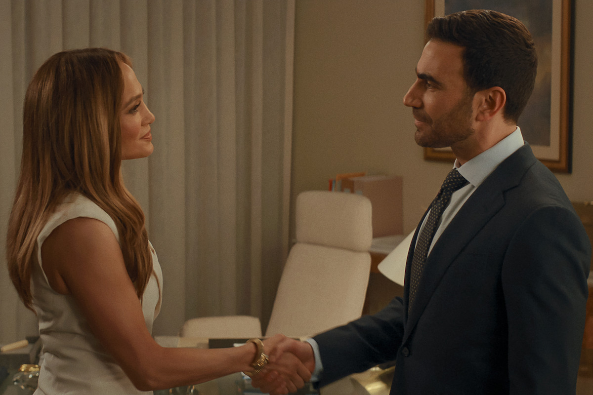 A woman in a white dress and a man in a suit are shaking hands in an office setting with a desk, chair, and soft lighting in the background.