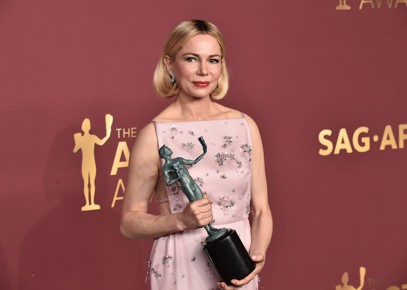 Michelle Williams in pink floral dress holding a trophy at an awards event, standing in front of a red backdrop with gold SAG Awards logos.