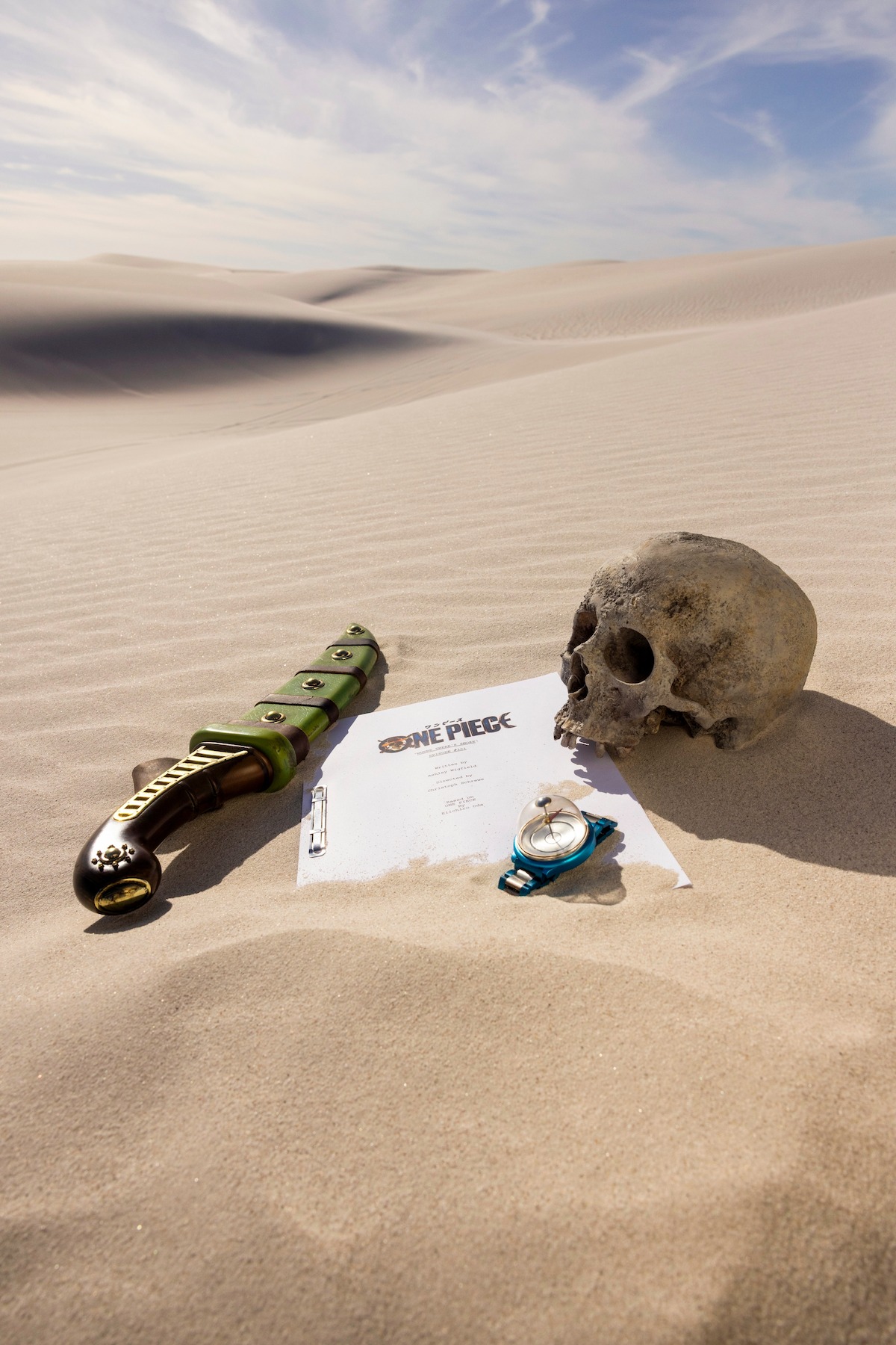 A sword, a human skull, a compass, and a ‘ONE PIECE’ script page arranged on desert sand, evoking an adventurous, mysterious, and slightly ominous atmosphere.
