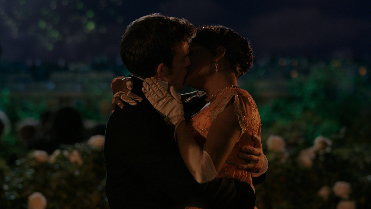 A couple dressed formally shares a passionate kiss at night in a beautiful garden surrounded by flowers and soft lights, suggesting a romantic and intimate atmosphere.