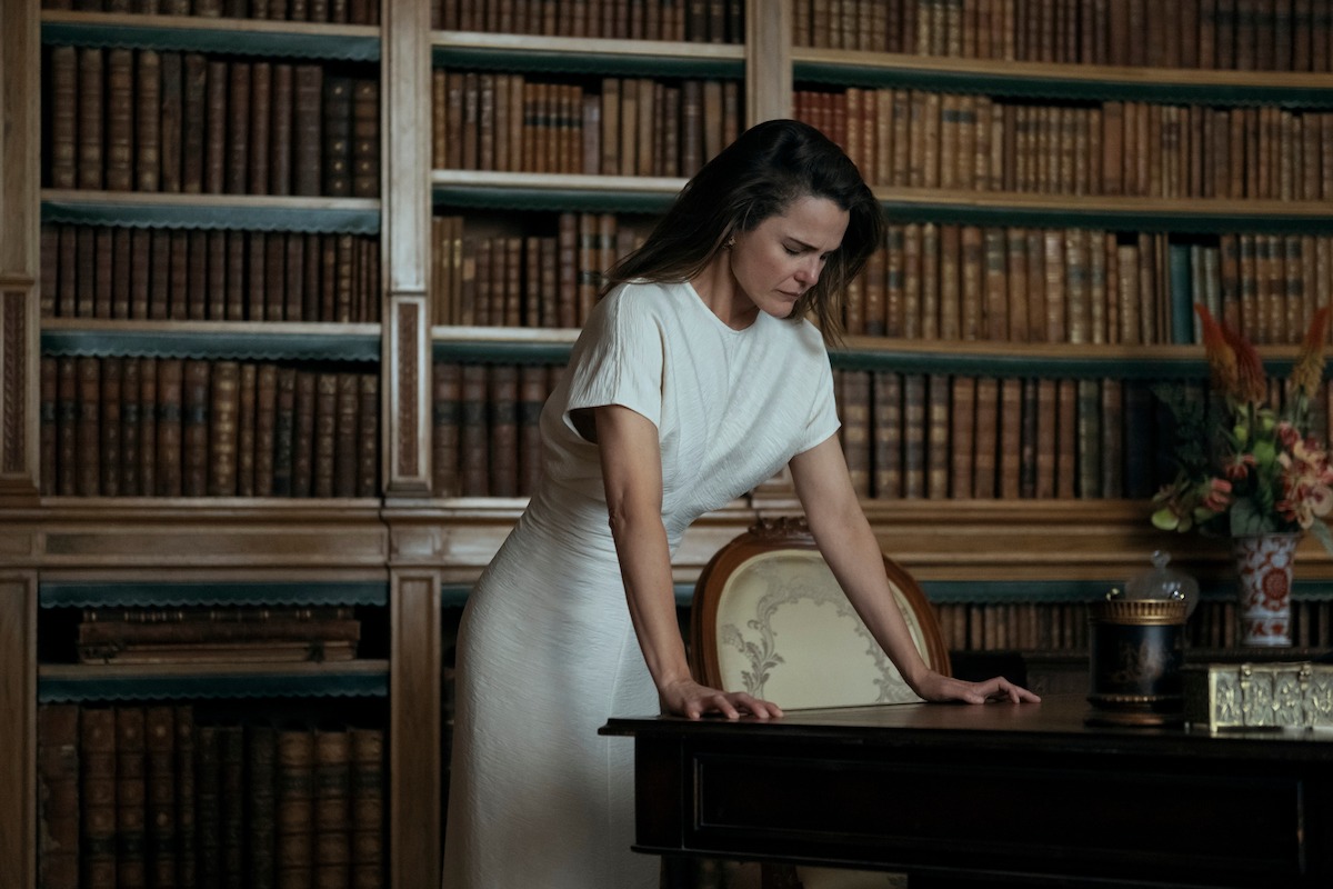 Woman in a white dress stands leaning on a desk in a library filled with bookshelves, appearing thoughtful or concerned; the room has classic wooden decor and a vase of flowers on the side.