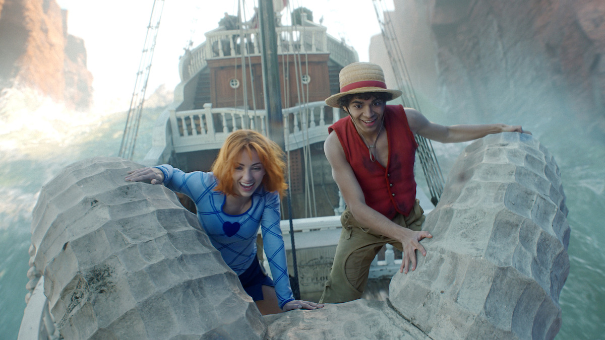 Two people on a ship’s figurehead, one with orange hair in a blue outfit and the other wearing a straw hat and red vest, smiling as the ship sails between cliffs and waves.